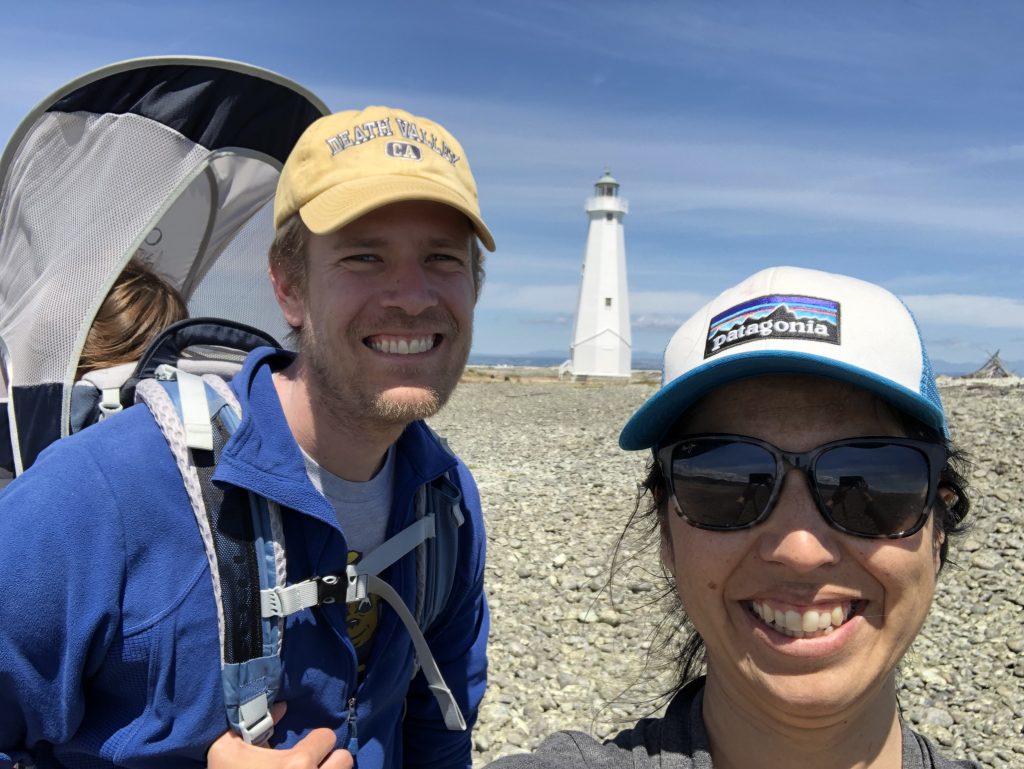 Two adults with a child asleep in an Osprey Poco in front of the Boulder Bank Lighthouse in Nelson NZ
